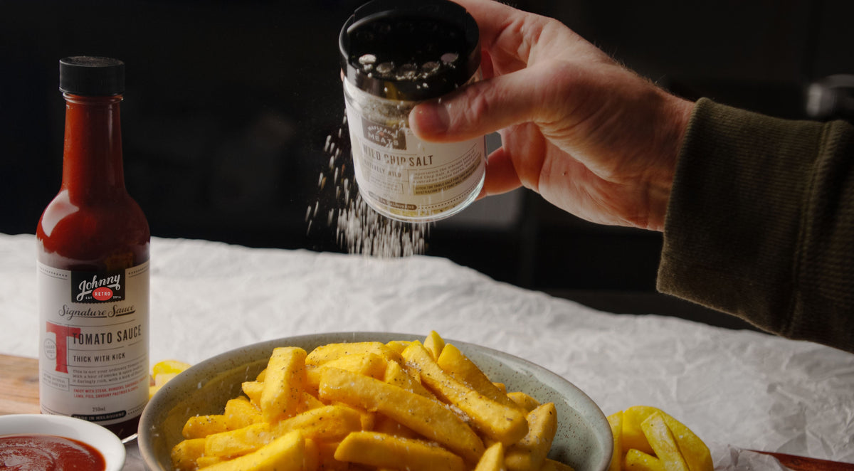 Person seasoning fries with Johnny Retro Wild Chip Salt, with Johnny Retro tomato sauce on a table.